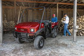 A red Kawasaki Mule utility vehicle in a shed with two men loading chopped wood in the back.