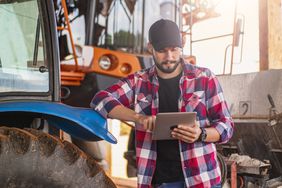 Man in a black hat and a red, blue, and white flannel leans against a tractor while scrolling on his tablet. 