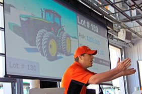 Auctioneer in a red shirt and hat begs bids for a green John Deere tractor. 