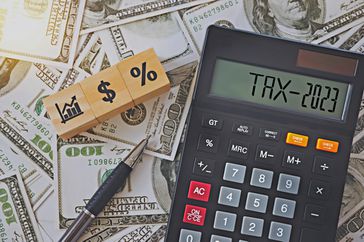 A black calculator reading "TAX-2023" sits next to a pen and building blocks with the images of a graph, a dollar sign, and a percent sign, all on a background of U.S. $100 bills