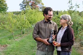 Man and woman stand in an orchard, holding a bottle of cider.