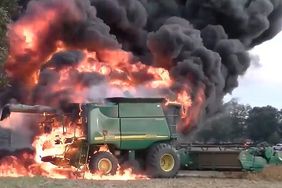 A John Deere combine engulfed in flames sits in a soybean field with black smoke billowing