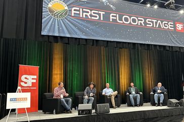 Four men sit on a stage under a "first floor stage" banner