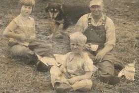 Three people sit in a field with a black dog 