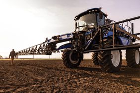 The New Holland Guardian Sprayer with boom arms extended in a field at sunset.