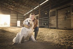 A woman in chore boots and a coat kneels next to a large guard dog inside a horse barn