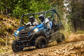 A blue Kawasaki Teryx side-by-side with two passengers riding off-road in the woods.