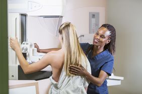 A mid-adult blonde woman getting a mammogram. She is being helped by a Black nurse. 