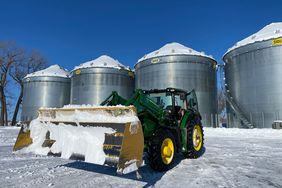 A John Deere tractor with a big scoop moves snow in front of grain bins on a sunny winter day