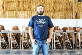 Oregon dairy farmer and author Derrick Josi stands in his cattle barn wearing a blue shirt and jeans